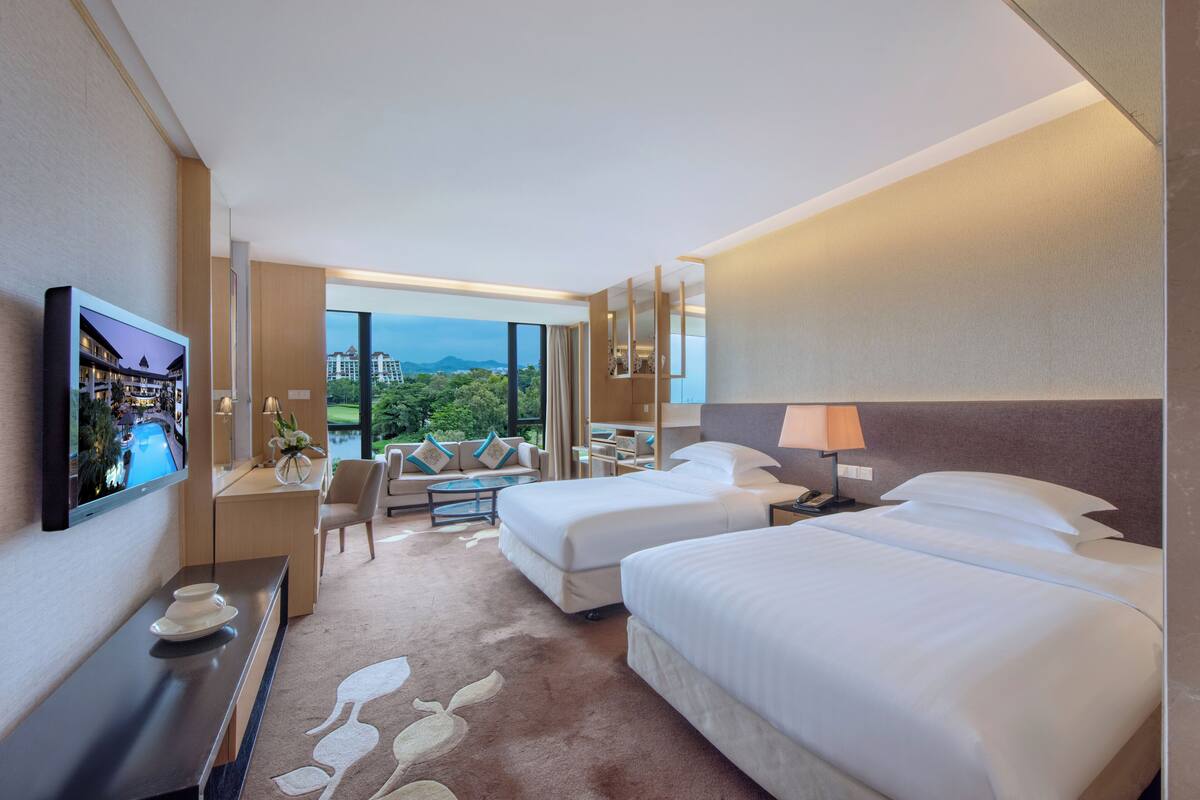 Ein Hotelzimmer mit zwei Betten, einem Schreibtisch, einem Fernseher und Blick auf eine Stadtlandschaft.