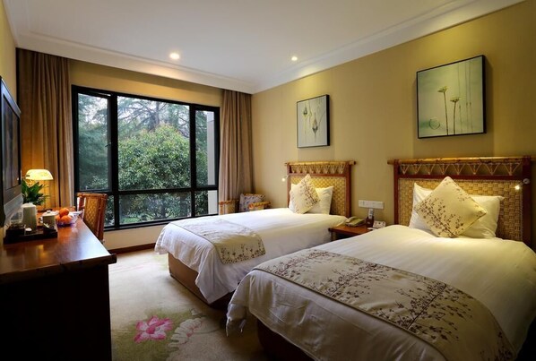 A hotel room with two beds, a desk with a lamp, and a large window offering a view of trees.