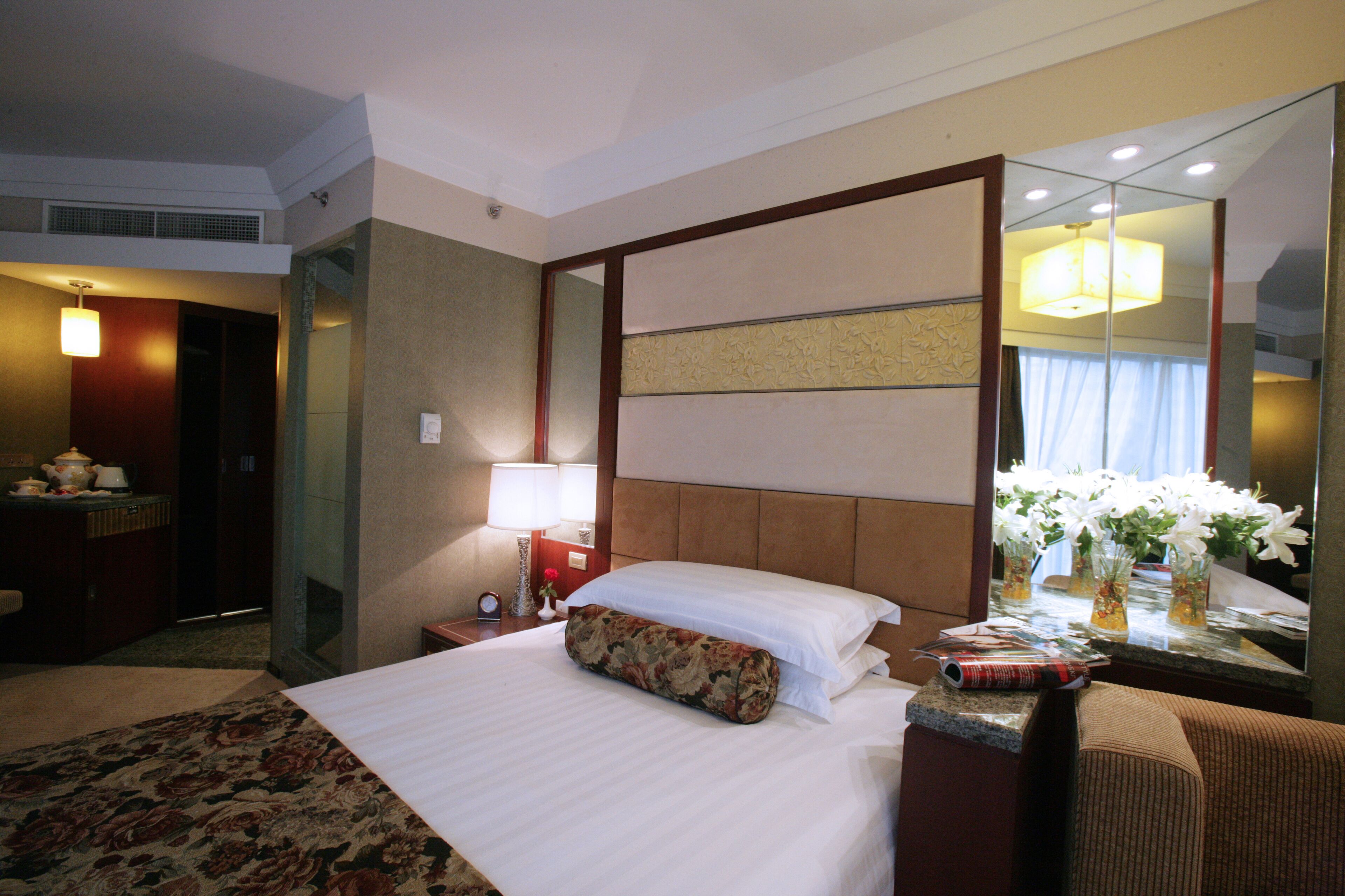 A hotel room with a large bed, a nightstand with a lamp, a mirror, and a floral-patterned pillow.