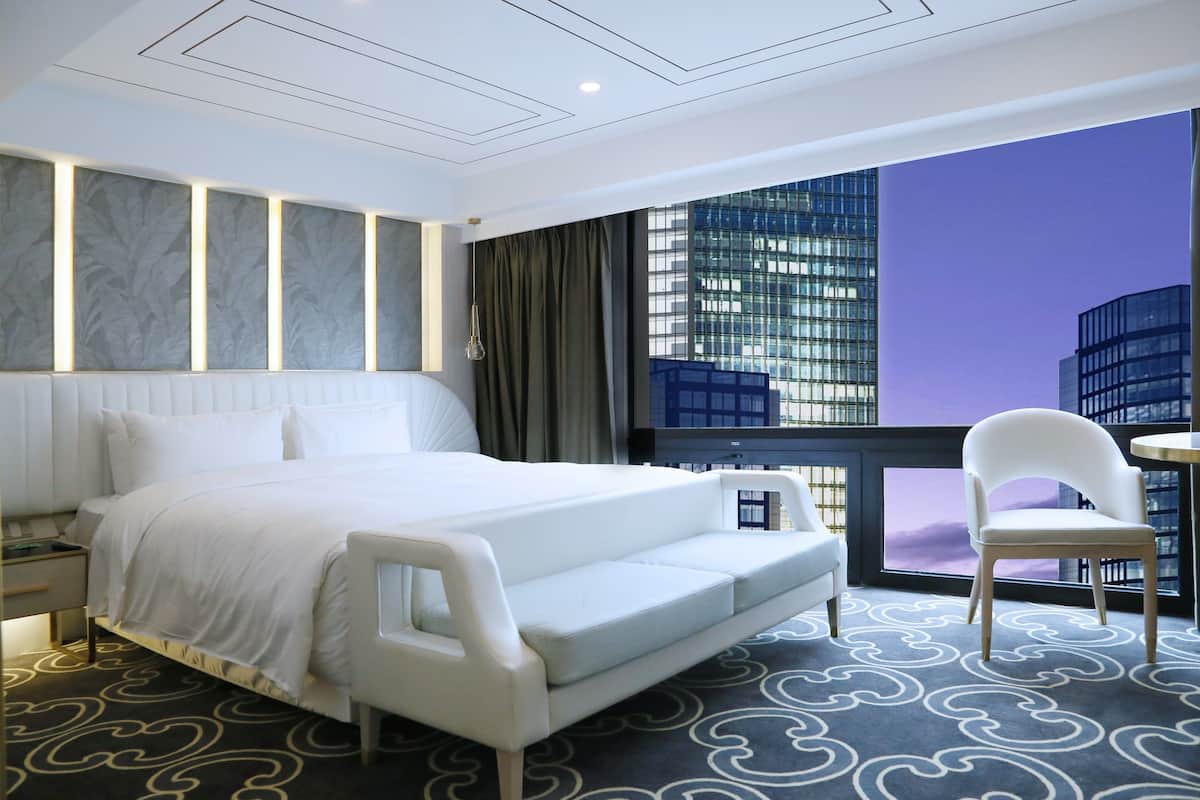 A modern hotel room with a large bed, a sofa, a chair, and a view of city buildings.