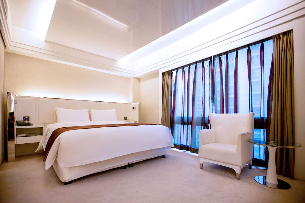 A modern hotel room with a large bed, a white armchair, a glass coffee table, and a view of the city through large windows.