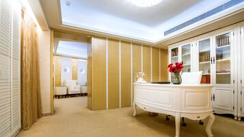 Vestíbulo de hotel con mostrador de recepción blanco, un área de descanso con dos sillas, una estantería y un jarrón decorativo con flores rojas.
