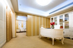 Vestíbulo de hotel con mostrador de recepción blanco, un área de descanso con dos sillas, una estantería y un jarrón decorativo con flores rojas.