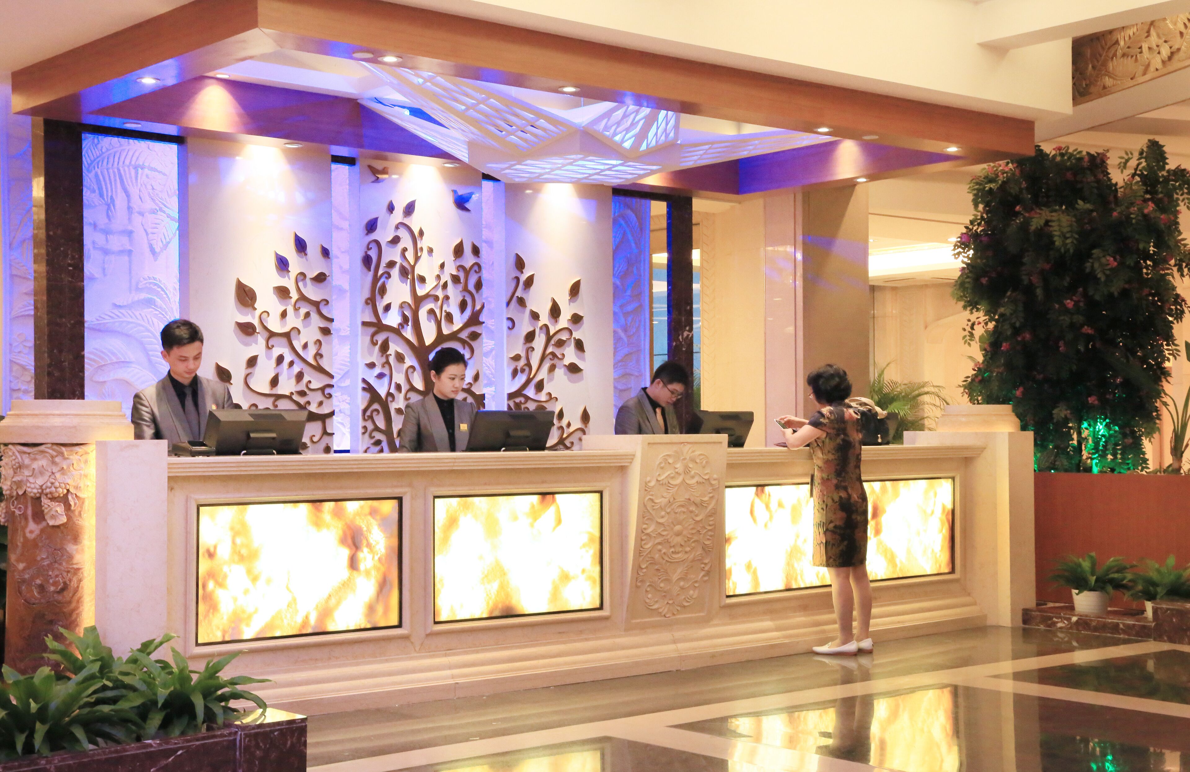 A hotel reception area with a modern design, featuring a reception desk with multiple screens displaying a fire animation, decorative wall panels, and potted plants.