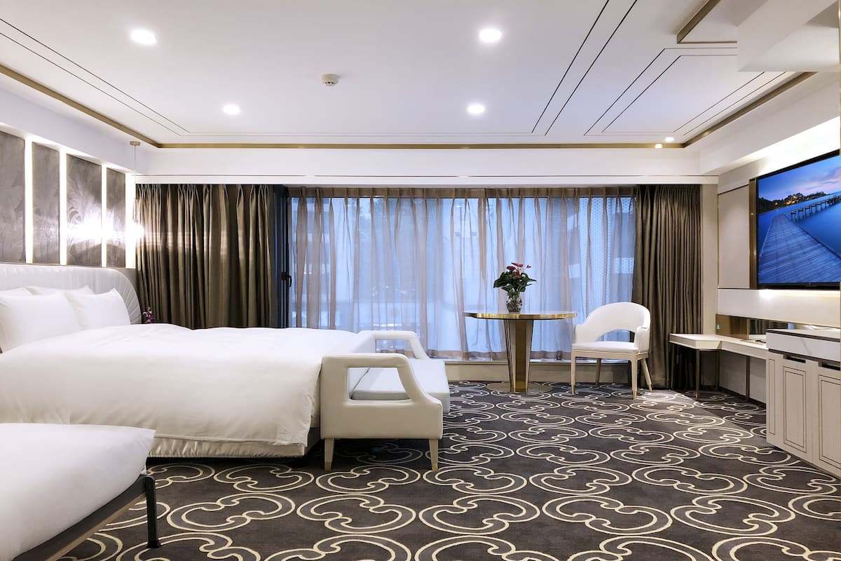 A hotel room with a large bed, two chairs, a small table with a vase, and a TV on the right.