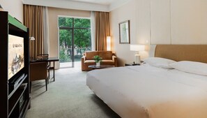 A hotel room with a bed, a desk, a chair, a TV, and a view of greenery outside.