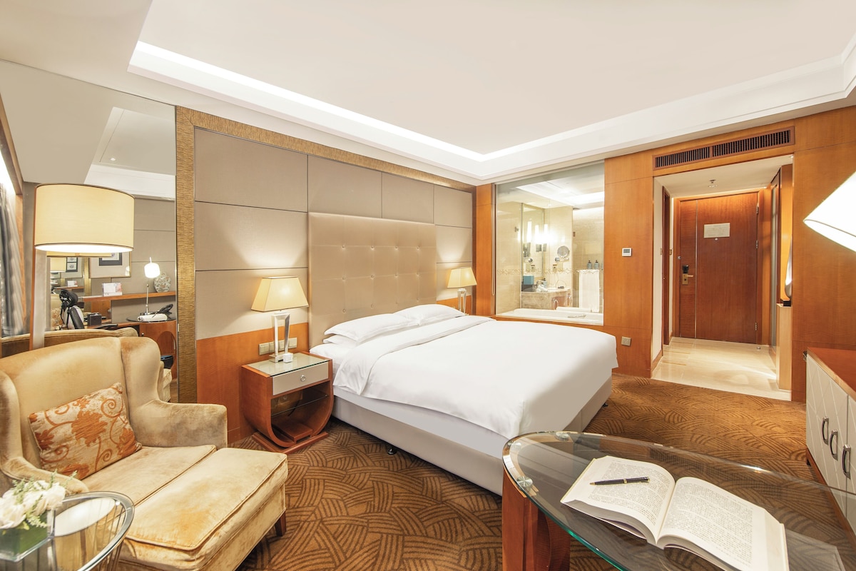 A hotel room with a bed, a sofa, a glass table with an open book, a nightstand with a lamp, and a mirror.
