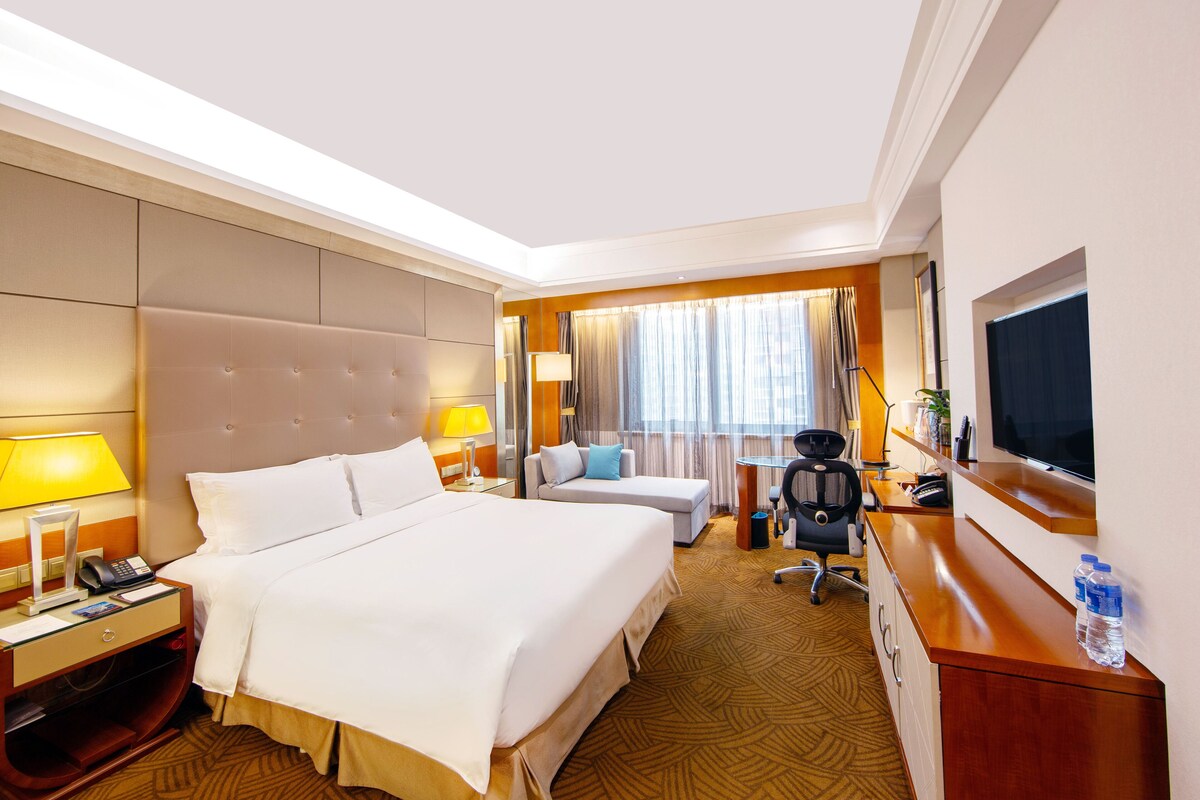 A hotel room with a large bed, a desk with a chair, a TV, and a desk lamp.