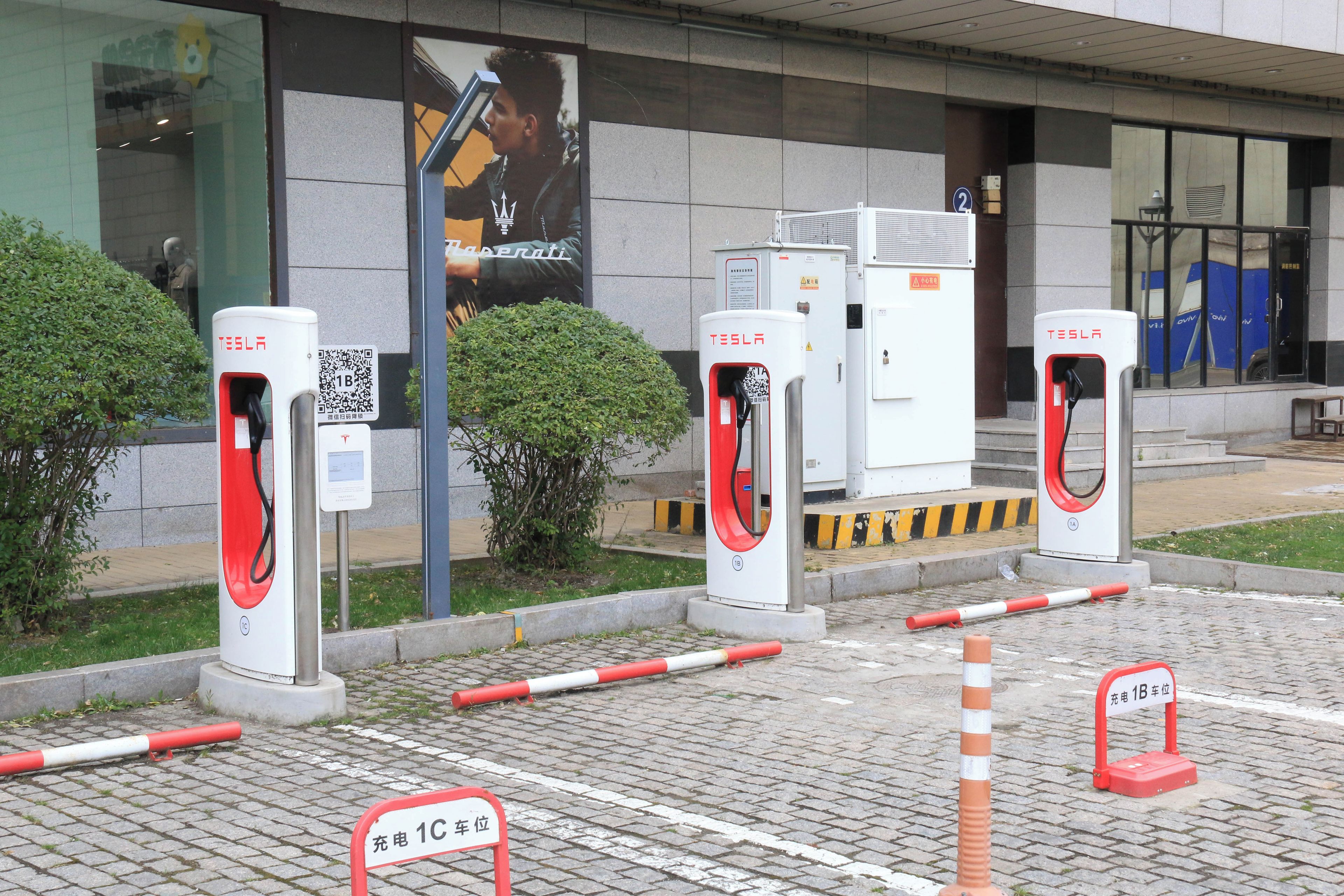 Un área de estacionamiento con varias estaciones de carga para vehículos eléctricos, la entrada de un edificio y un letrero que indica la ubicación de las estaciones de carga.