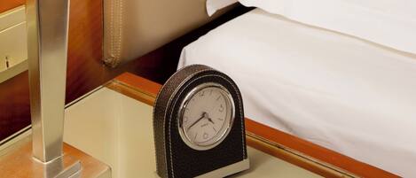 A neatly made bed with white sheets, a bedside lamp, and a classic analog clock on a bedside table.