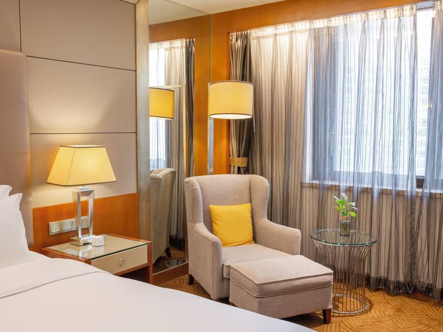 Een hotelkamer met een bed, een stoel met een geel kussen, een glazen tafel met een vaas en gordijnen voor de ramen.