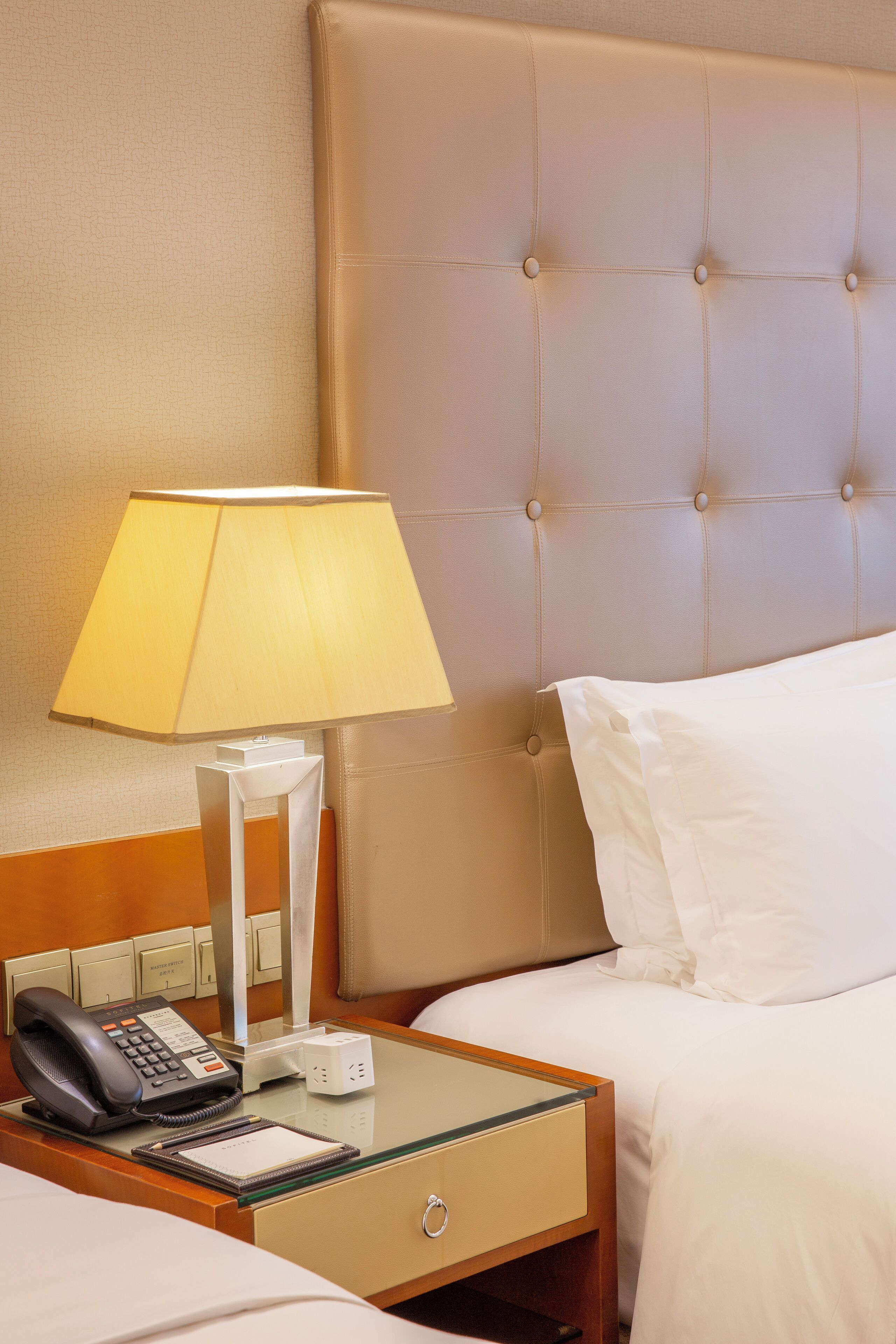 Habitación de hotel con cama, mesita de noche con lámpara, teléfono y un pequeño escritorio con bloc de notas.