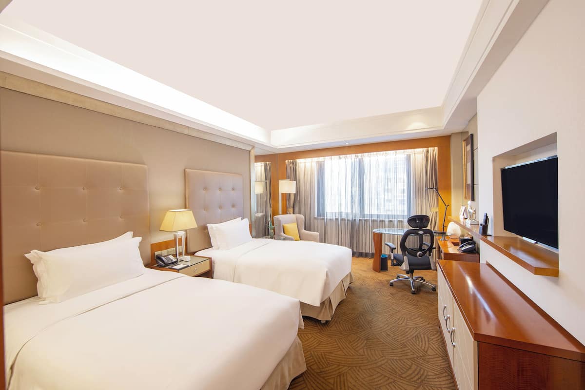 A hotel room with two beds, a desk with a chair, a television, and a window with curtains.