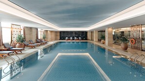 A large indoor swimming pool with lounge chairs and potted plants, surrounded by walls and a ceiling with recessed lighting.