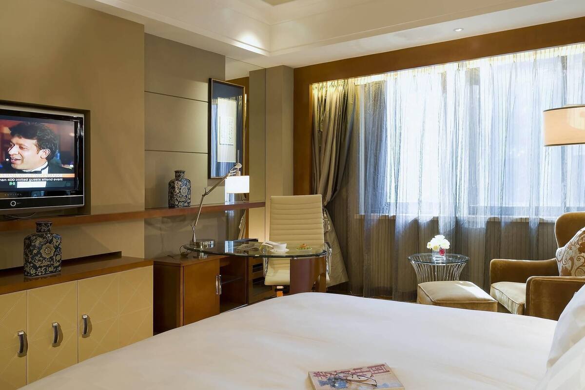 A hotel room with a bed, a desk, a television, a chair, a small table, and a window with curtains.