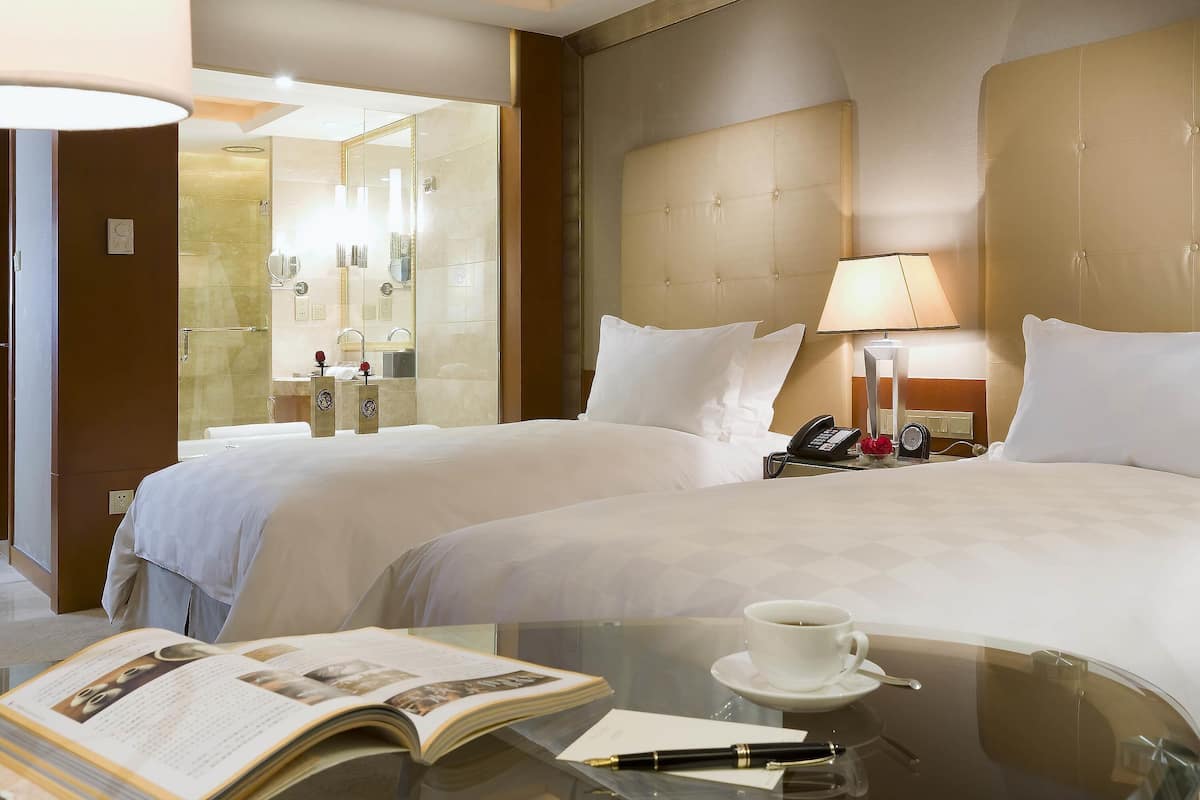 A hotel room with two beds, a glass coffee table with a book, a cup of coffee, and a bedside table with a lamp and a phone.