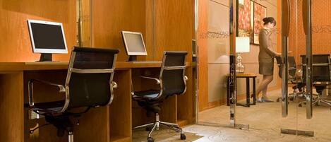 Zona de recepción de hotel con paneles de madera, dos monitores de computadora, sillas de oficina negras y una persona de pie junto a una puerta de vidrio.