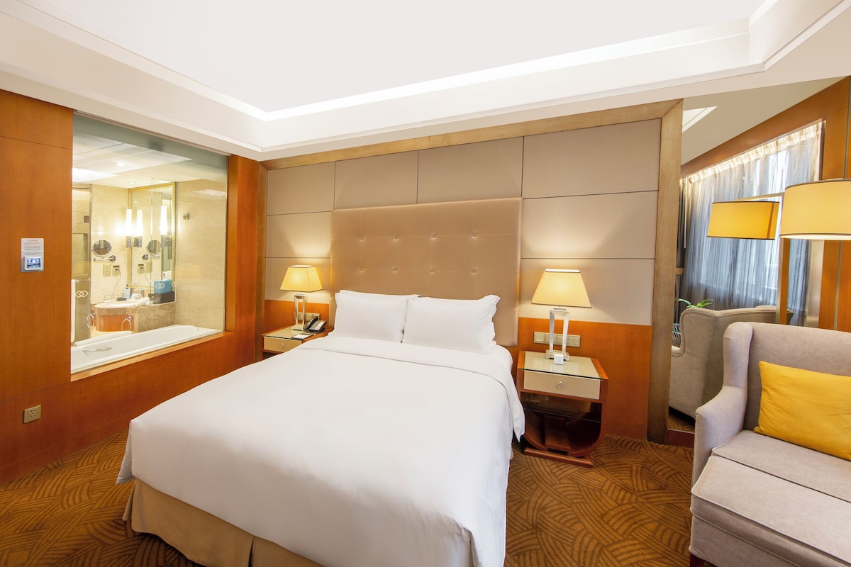 A hotel room with a large bed, two bedside tables with lamps, a chair, a mirror, and a window with curtains.