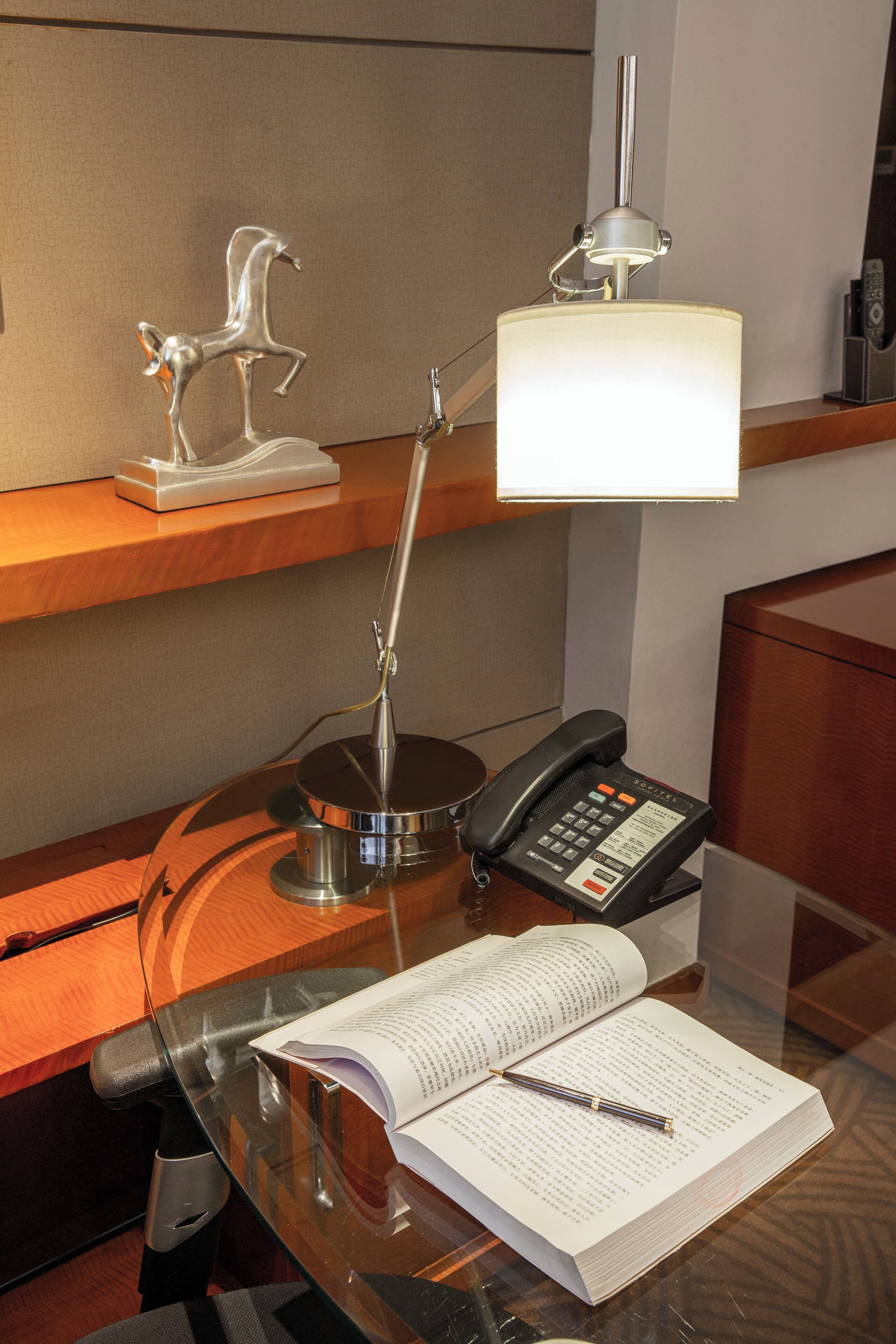 Ett skrivbord i ett hotellrum med en lampa, en telefon och en öppen bok.