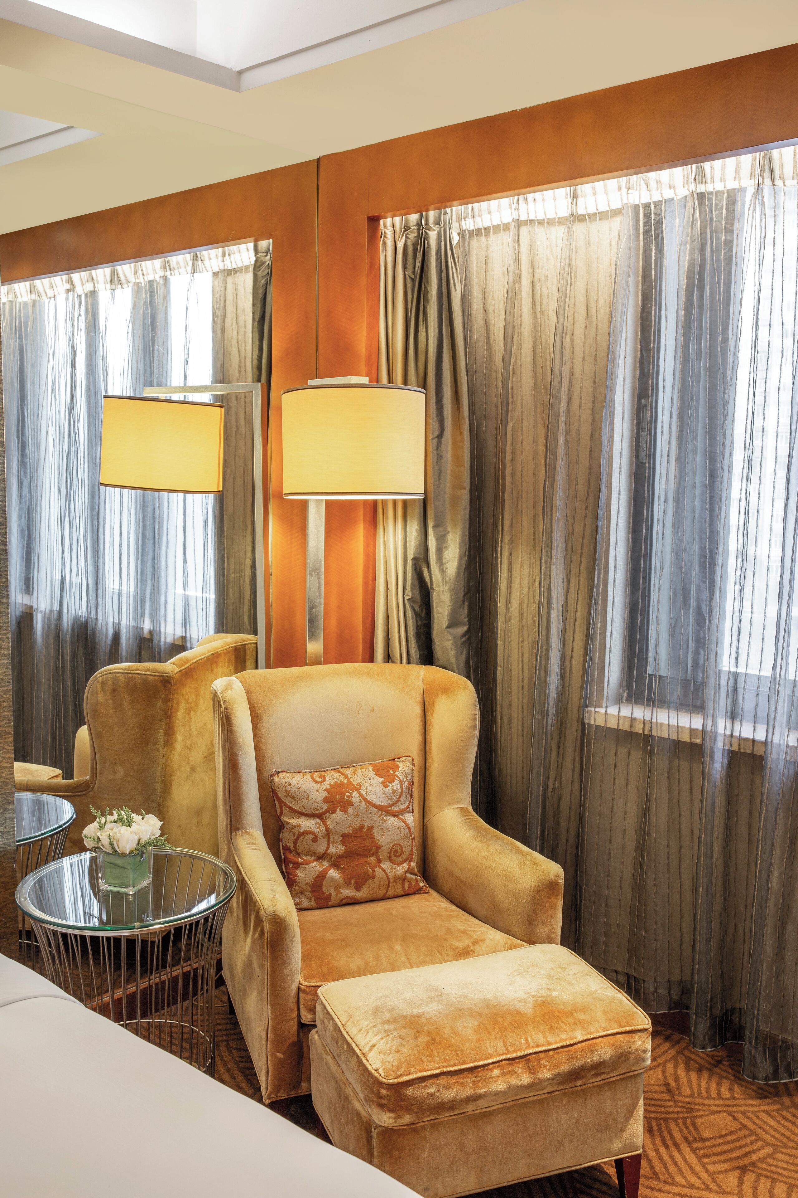 Una habitación de hotel con un sillón cómodo, una mesita redonda con un florero y un ventanal con cortinas translúcidas.