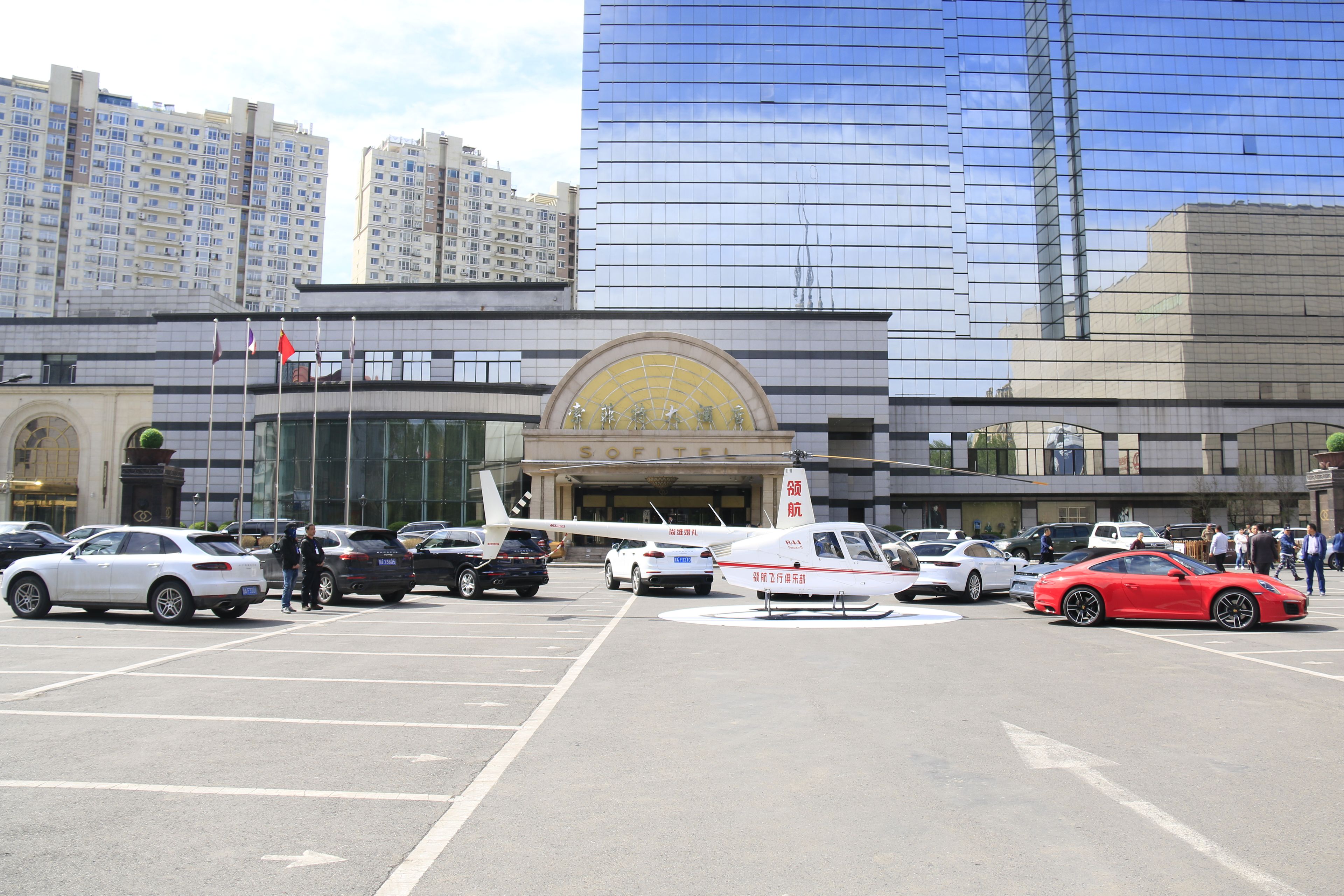 A parking lot with various cars, a helicopter, and a building with a large clock and signage in the background.