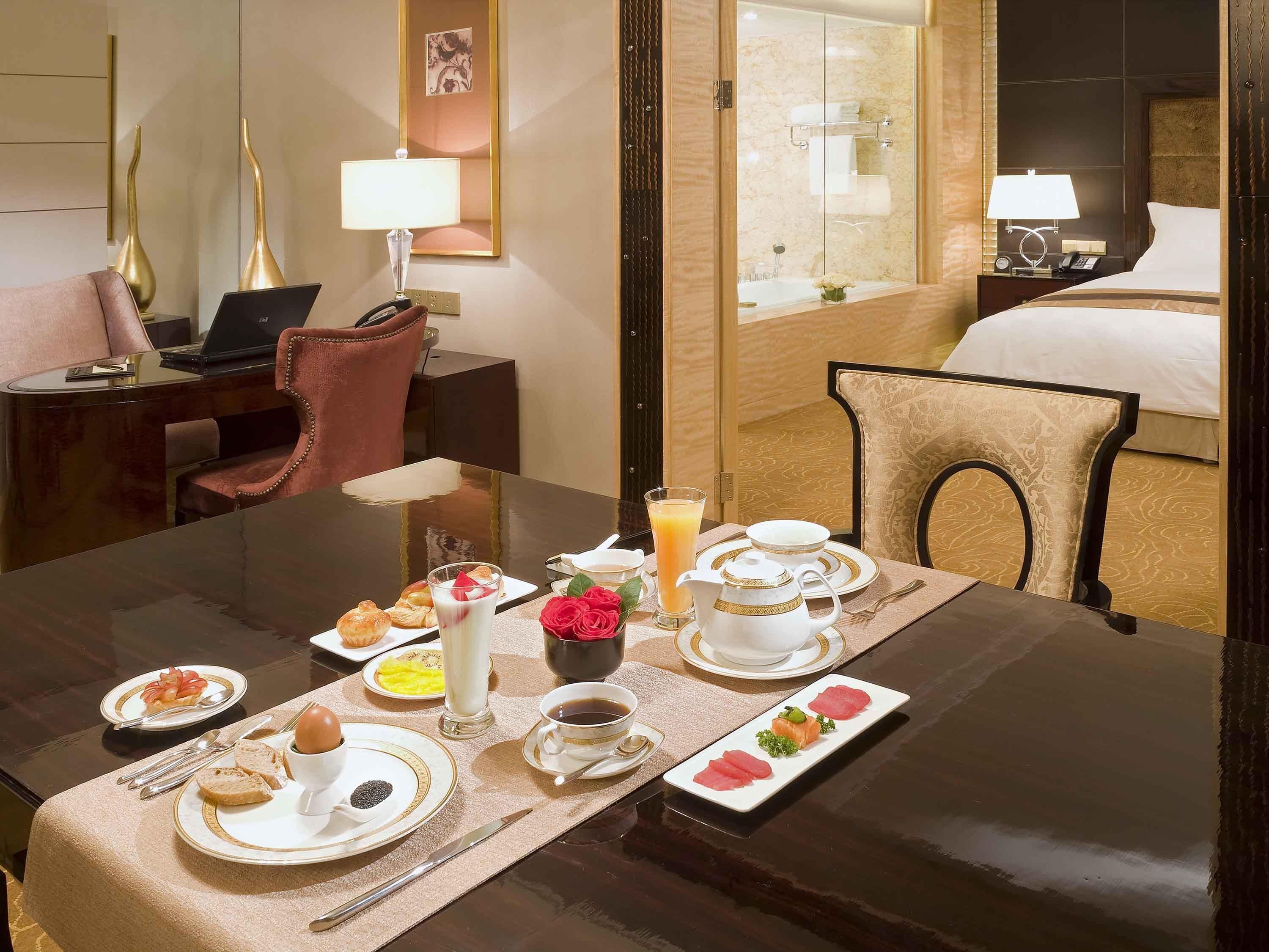 A hotel room with a dining table set for breakfast, a bed, a desk with a laptop, and a mirror.