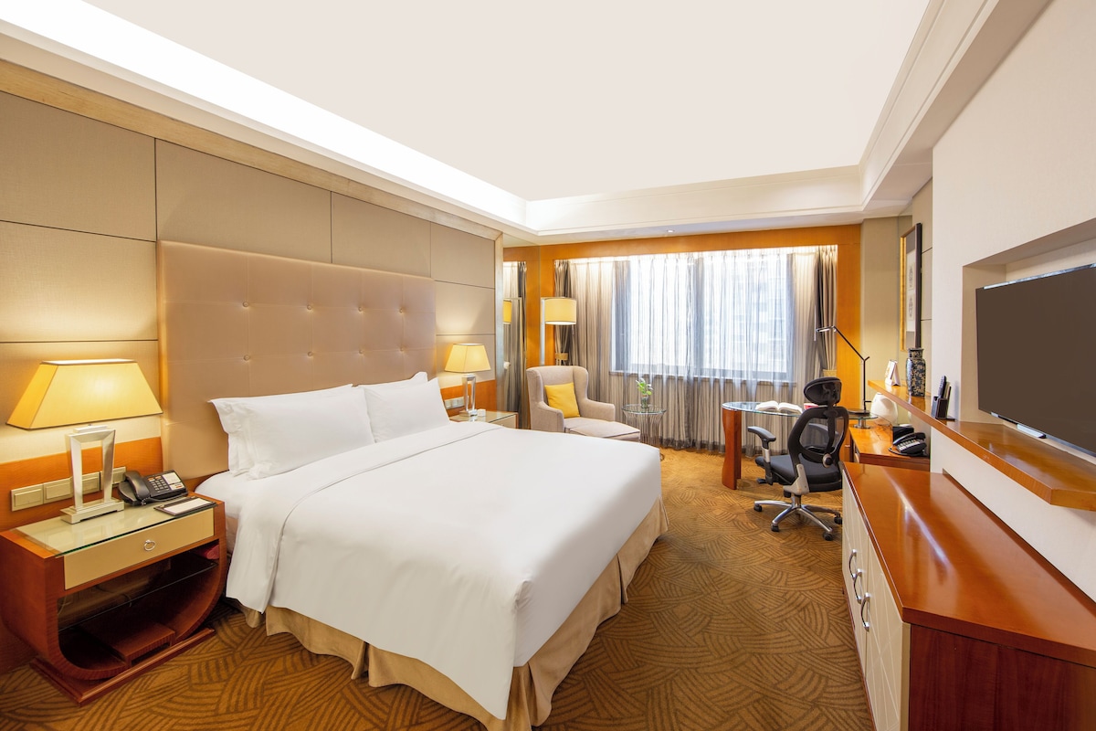 A hotel room with a large bed, bedside tables, a desk with a chair, a television, and a window with curtains.