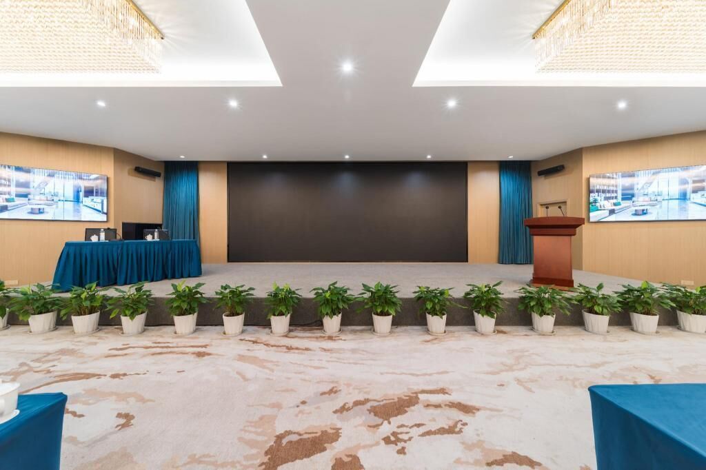 Un salón de conferencias con un escenario, un atril y una fila de plantas en macetas.