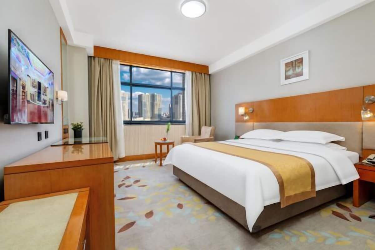 Una habitación de hotel con cama, escritorio, silla, televisión, ventana con vistas a la ciudad y un cuadro en la pared.