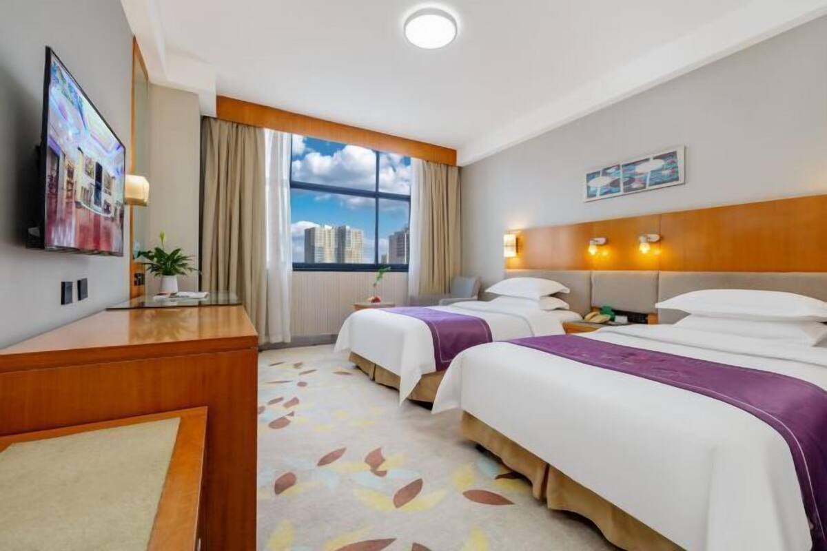 Habitación de hotel con dos camas, un escritorio, un televisor, una ventana con vista a la ciudad y un cabecero con luces.