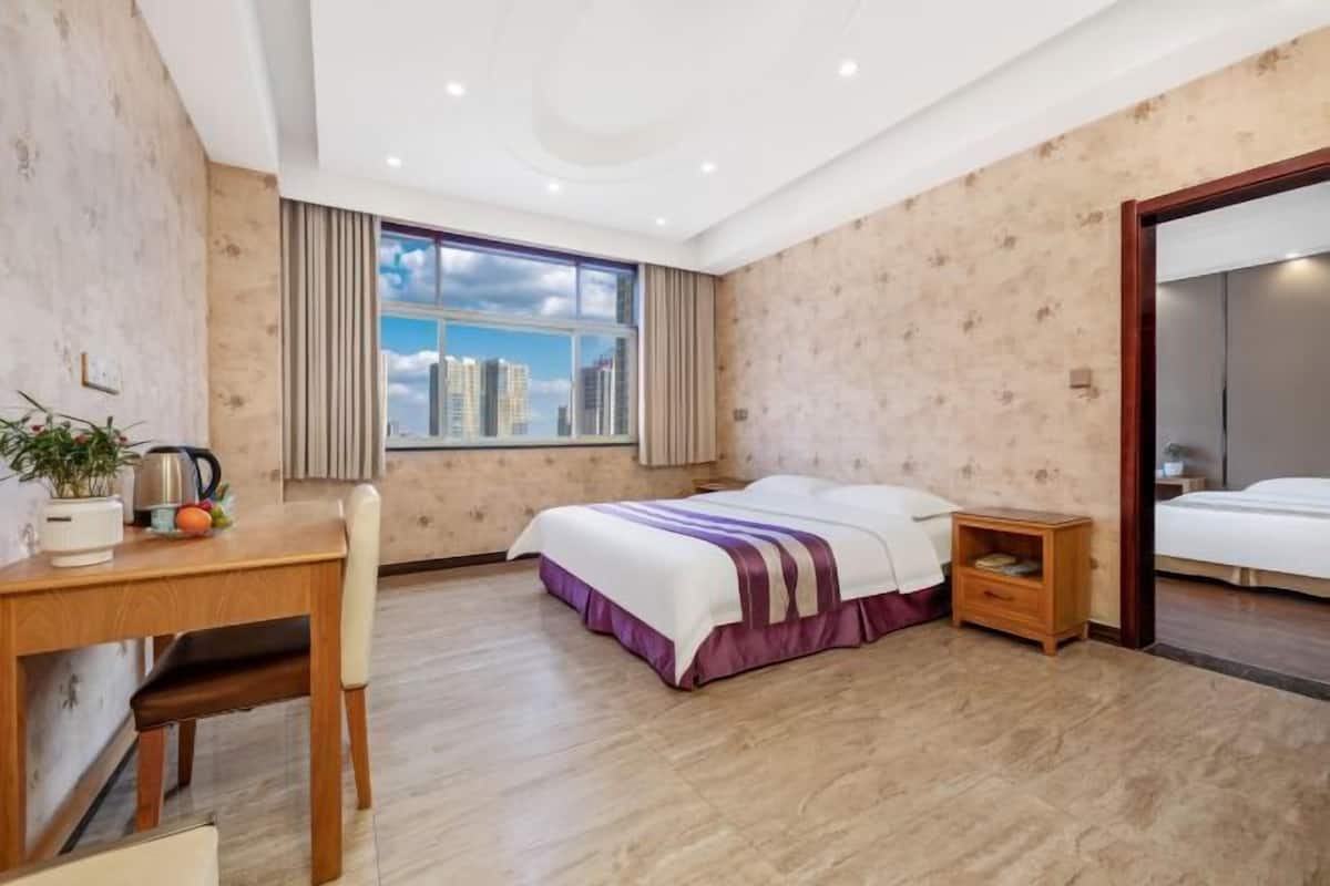 Habitación de hotel con cama, escritorio con silla, mesita de noche pequeña y ventana grande con vista a la ciudad.