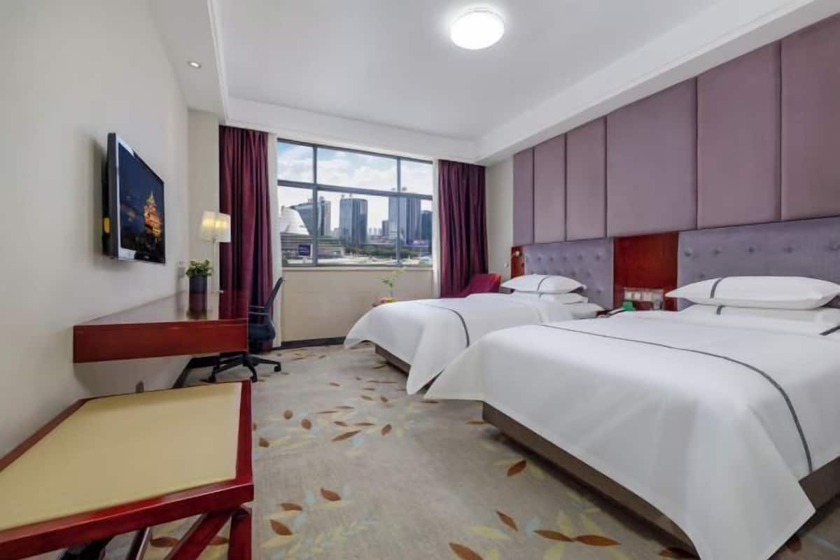 Habitación de hotel con dos camas, un escritorio, una silla, un televisor y ventana con vista a la ciudad.