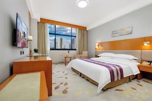 Habitación de hotel con cama, escritorio, silla, televisión, ventana con vista a la ciudad y un cuadro en la pared.