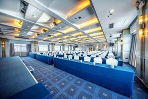 Une grande salle de conférence avec des rangées de tables et de chaises, un sol recouvert de moquette et un plafond haut doté d’un éclairage encastré.