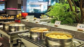A buffet area with various food items in stainless steel containers, a red hanging light fixture, and a large indoor plant. - Zhongdu Hotel (Zhengzhou)
