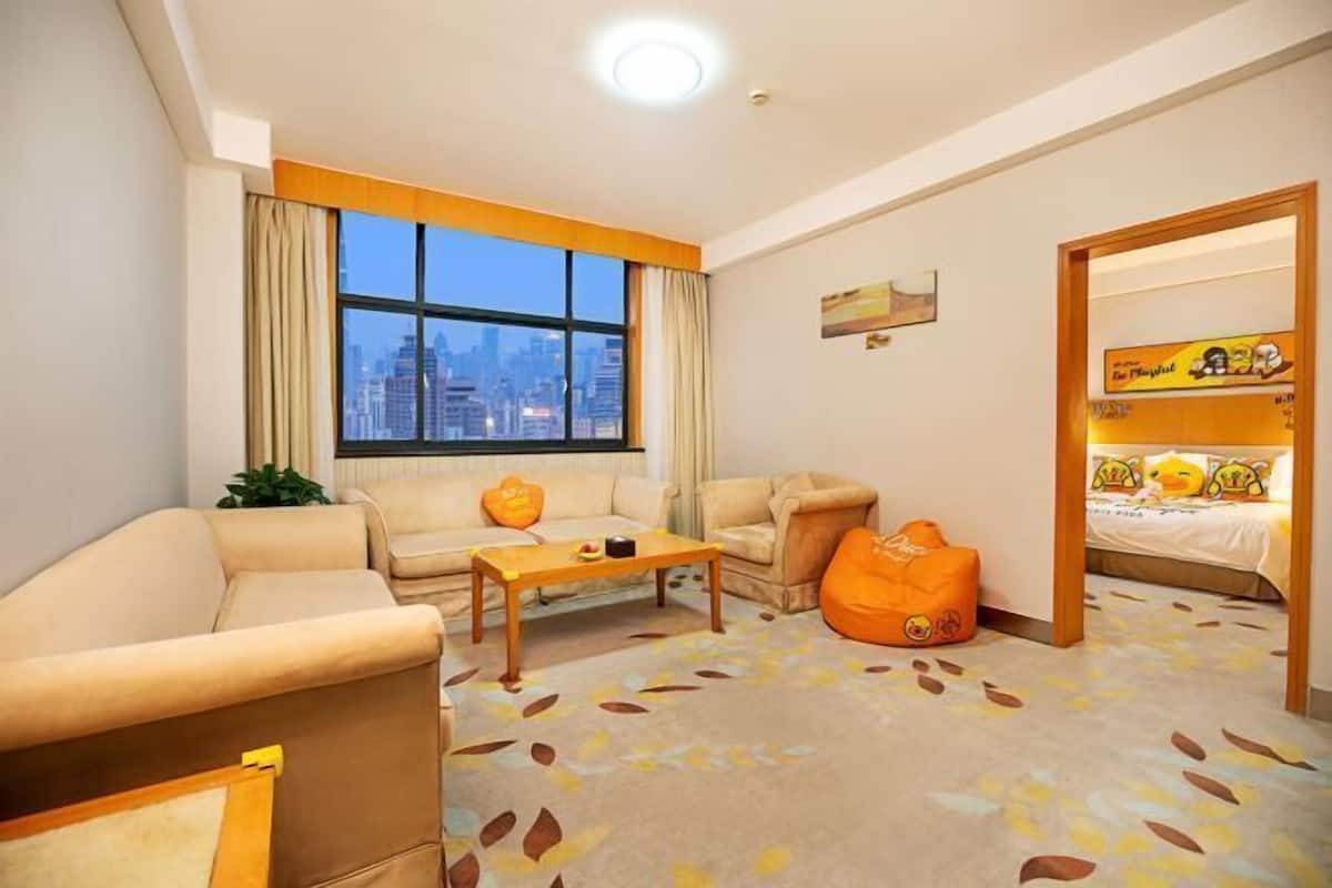 Una habitación de hotel con sofá, una mesita, un sillón de peluche, cama, ventana con vistas a la ciudad y puerta que da a otra habitación.