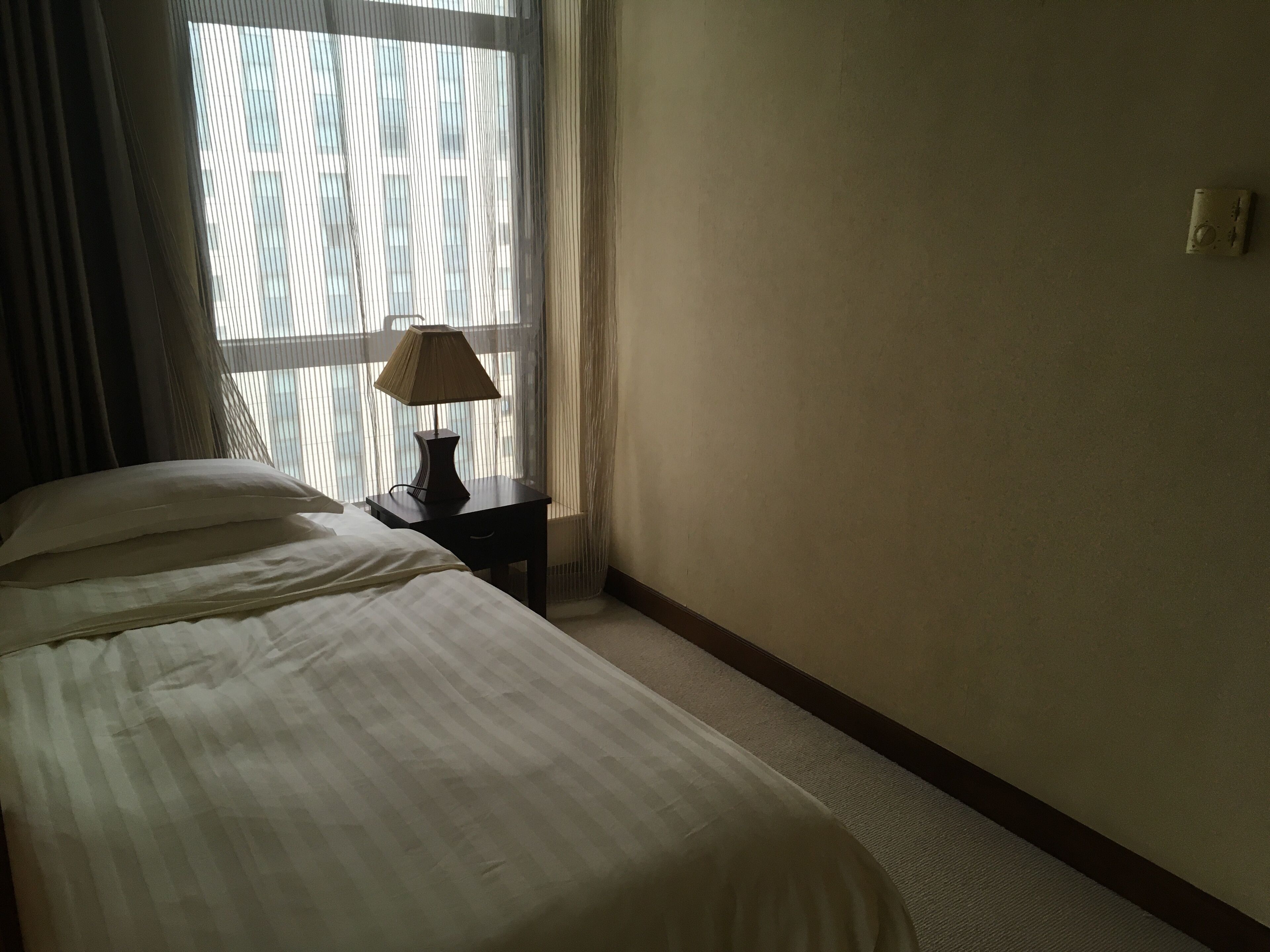 Une chambre d’hôtel avec un lit, une table de chevet avec une lampe et une fenêtre avec des rideaux légers.