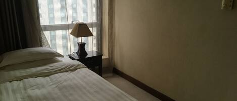 A hotel room with a bed, a nightstand with a lamp, and a window with sheer curtains.