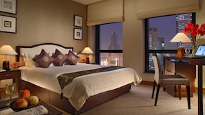 A hotel room with a large bed, two bedside tables, a desk with a chair, a view of the cityscape, and a window with a view of a tall building. - Dalian Asia Pacific Service Apartment (Dalian)
