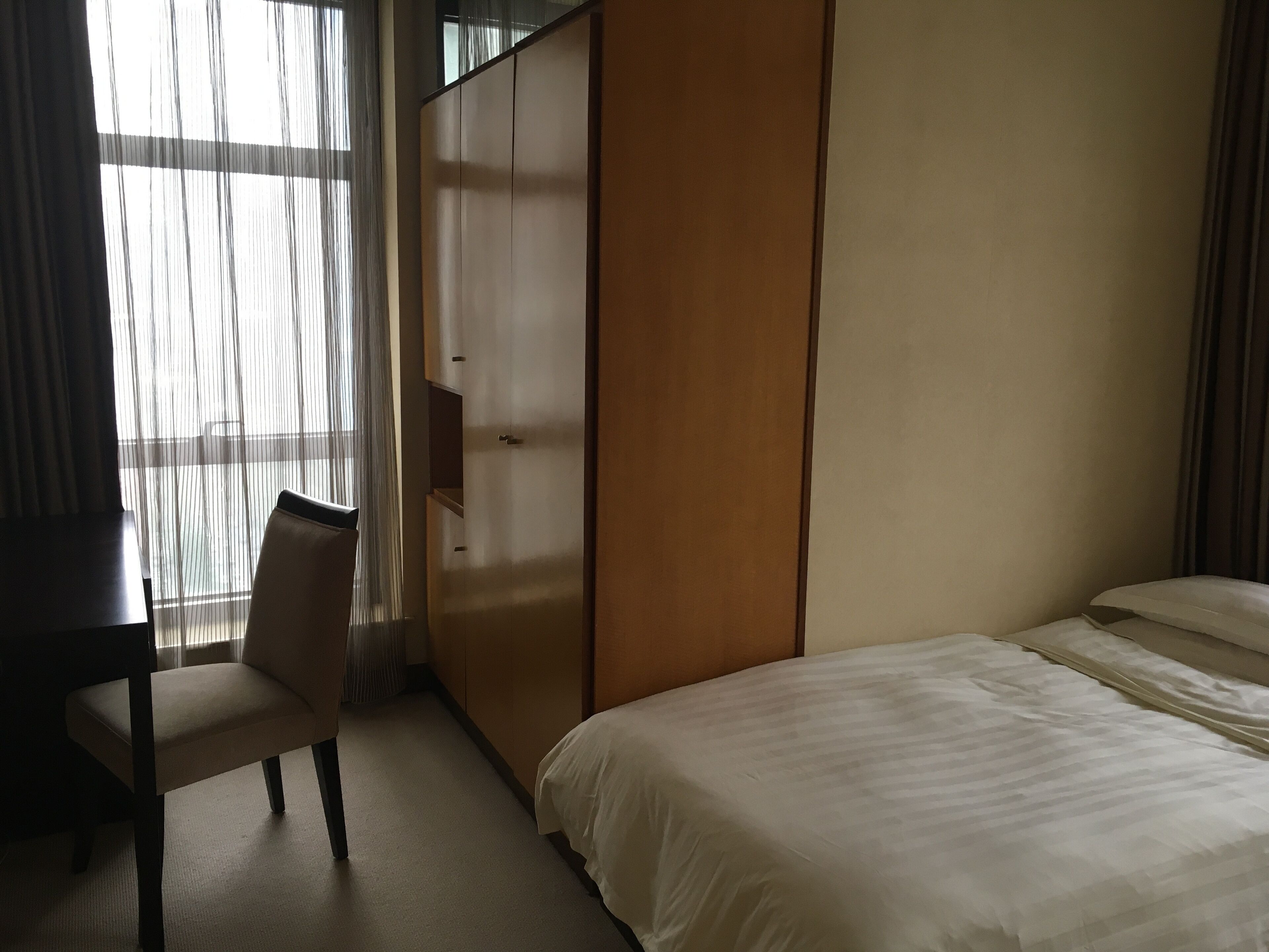 A hotel room with a bed, a chair, a wardrobe, and a window with sheer curtains.
