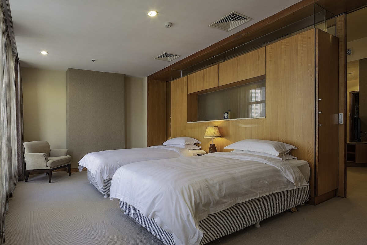 A hotel room with two beds, a chair, a lamp, and a large mirror.
