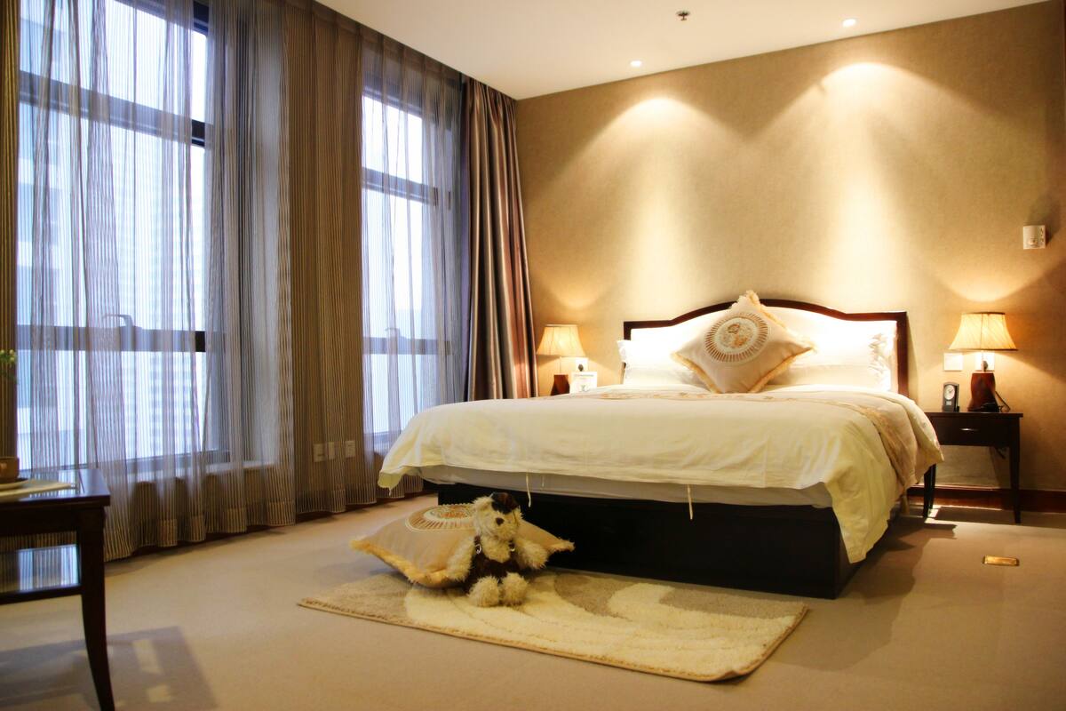 A hotel room with a large bed, two bedside lamps, a small table, and a teddy bear on a rug.