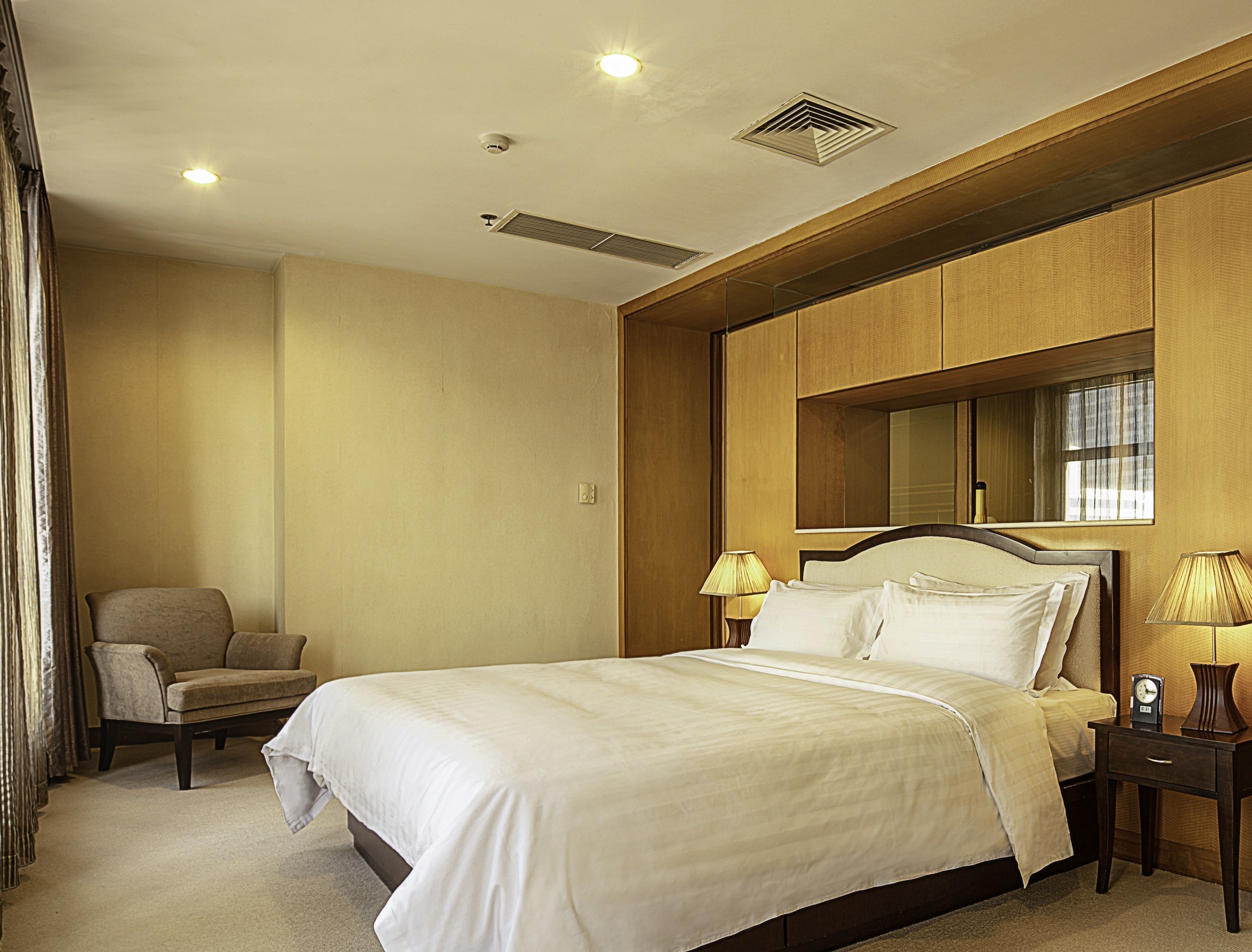 A hotel room with a bed, a chair, a nightstand with a lamp, and a large mirror on the wall.