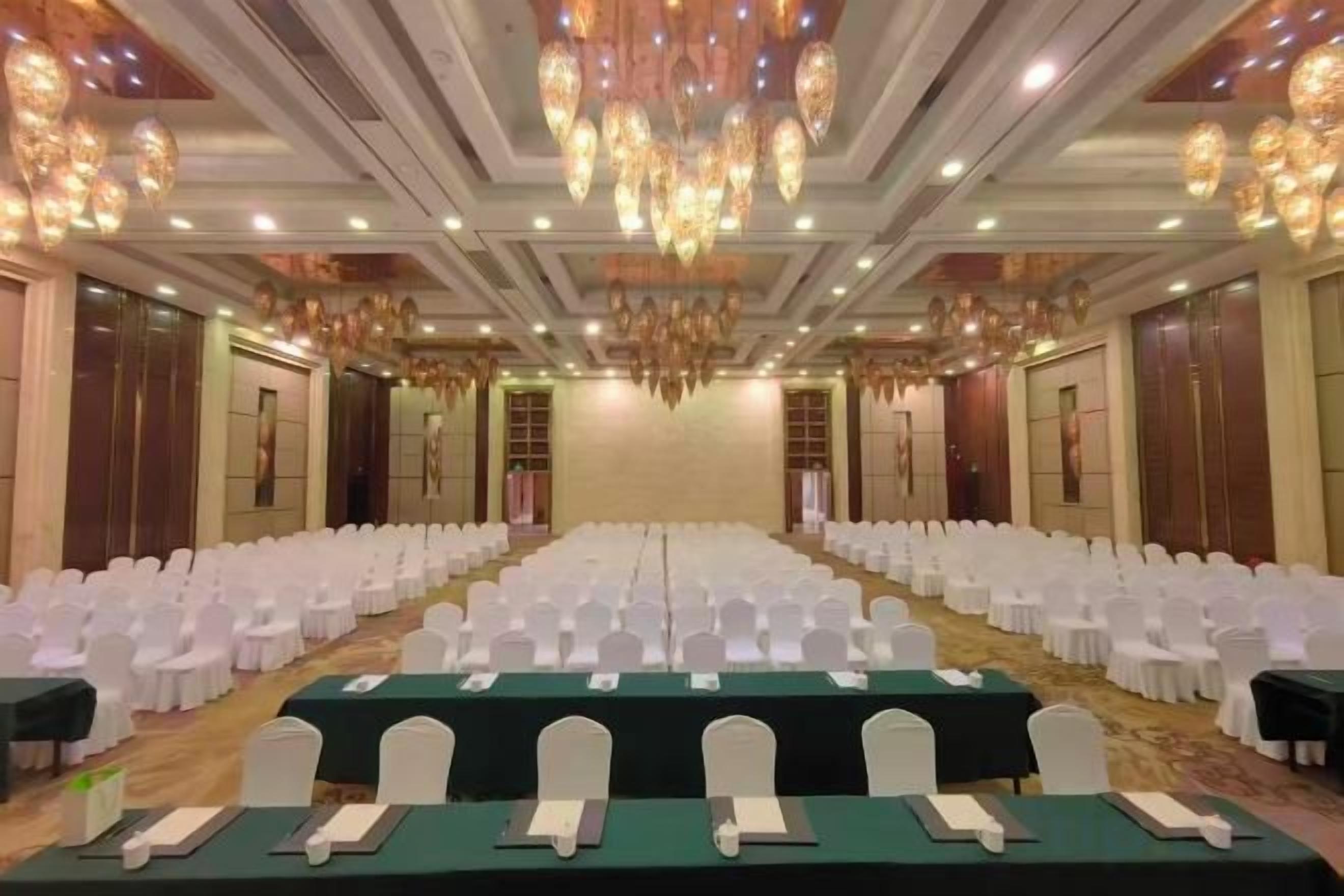 A banquet hall with rows of white chairs, tables with green tablecloths, and a chandelier.