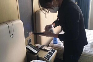 Una persona limpiando un ventanal en una habitación con cama, una mesita de noche con lámpara y panel de control de la ventana.