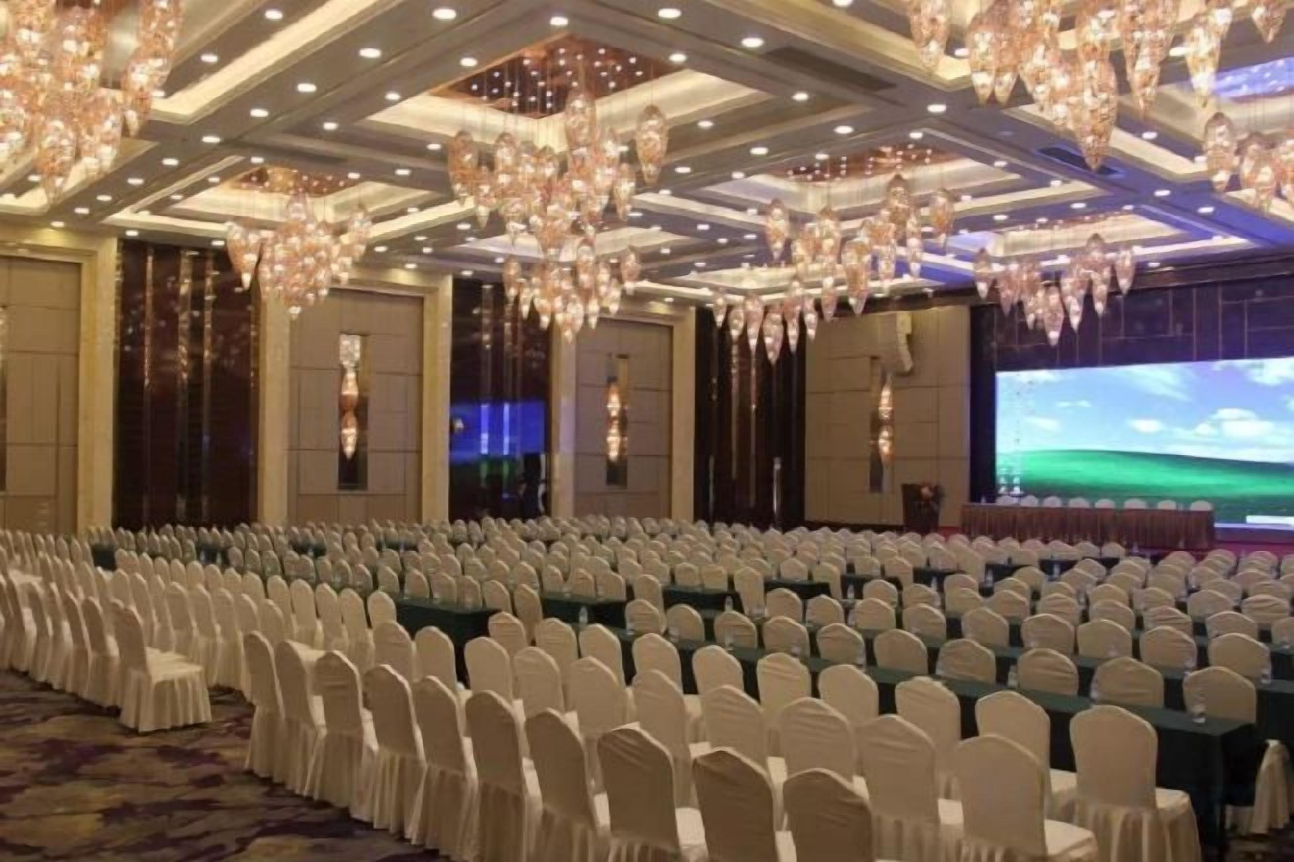 A banquet hall with rows of white chairs, a large screen, and chandeliers.