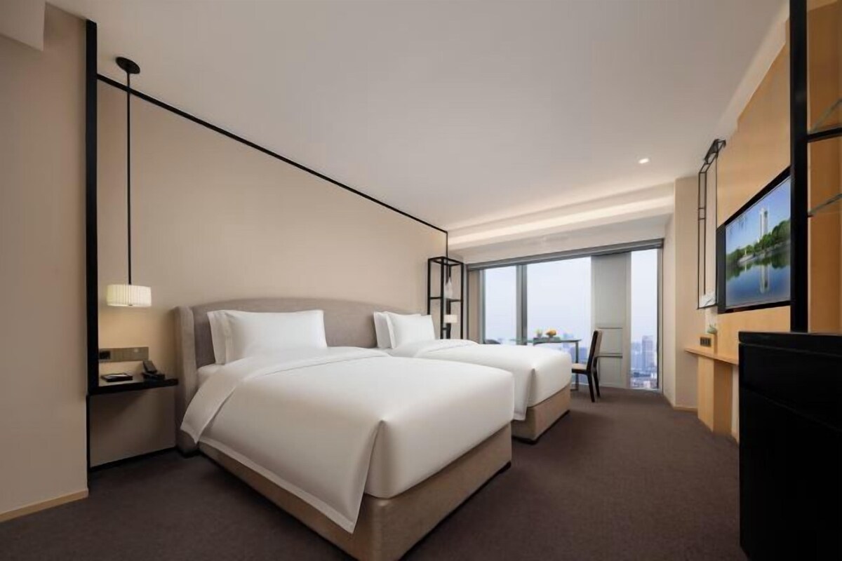 A hotel room with two beds, a TV, a desk, and a large window.