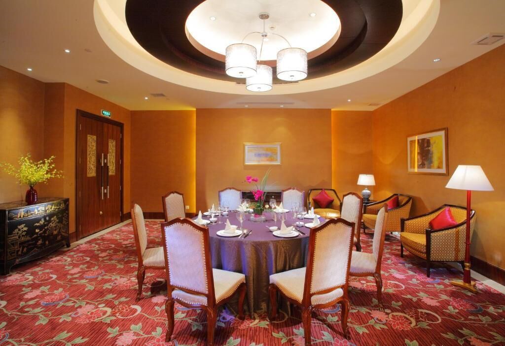 A conference room with a round table, four chairs, a chandelier, a floral carpet, and a cabinet with artwork.