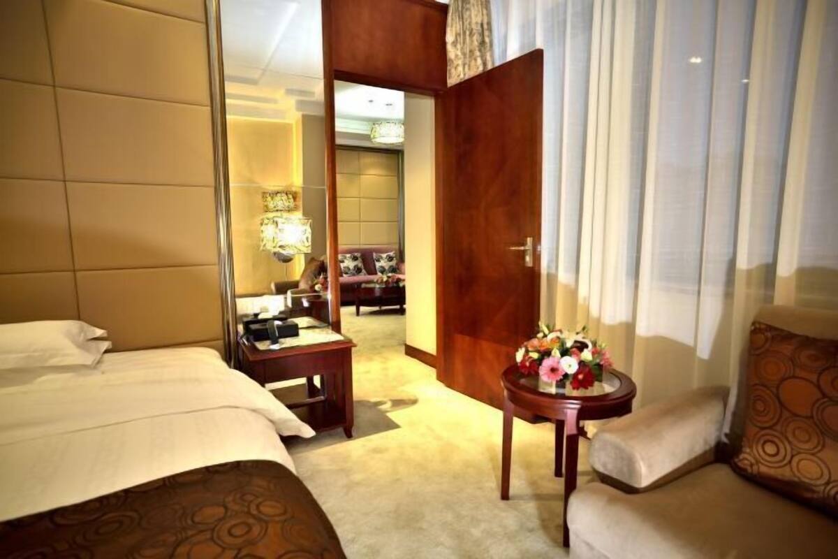 A hotel room with a bed, a small table with a vase of flowers, a sofa, and a view of a dining area.