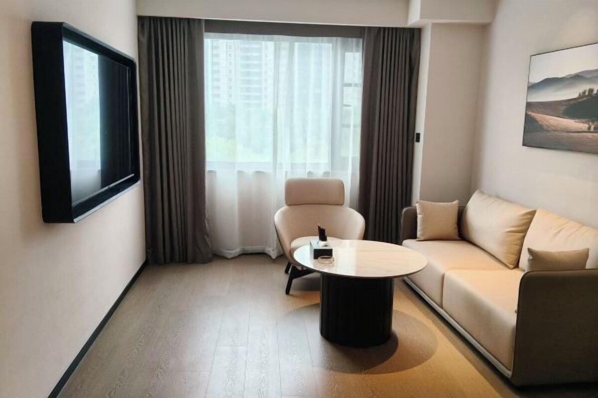 Una habitación de hotel moderna con sofá, una mesa redonda de centro, televisor montado en la pared y un ventanal con cortinas.